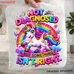 Puede incluir: Una hoja de tela blanca con un gráfico colorido de un unicornio con crin y cola de arcoíris, frente a un arcoíris. El texto dice NOT DIAGNOSED BUT SOMETHING ISN'T RIGHT. El diseño incluye nubes rosas, estrellas y corazones.