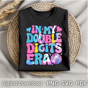 May include: Black t-shirt with the text "IN MY DOUBLE DIGITS ERA" in a colorful, retro font. The design includes hearts, leopard print, and a disco ball graphic. The shirt is displayed on a woven basket.