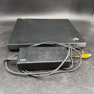 Puede incluir: Un adaptador de corriente IBM negro con un cable negro enrollado y un conector amarillo, colocado frente a una computadora portátil negra cerrada que presenta un pequeño logotipo multicolor en la superficie superior de la carcasa.