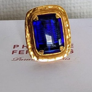 Könnte beinhalten: Ein goldener Ring mit einem großen, tiefblauen, rechteckigen Edelstein, der in einer strukturierten Goldfassung sitzt.