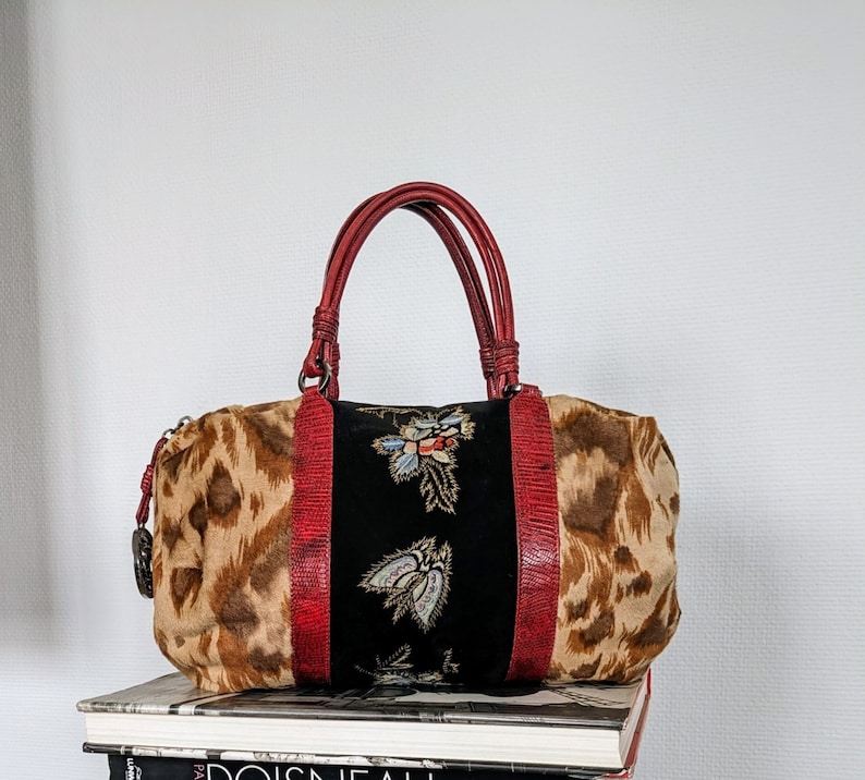 May include: A red, brown, and black leopard print handbag with a black velvet panel featuring embroidered floral designs. The bag has a top handle and a zipper closure.