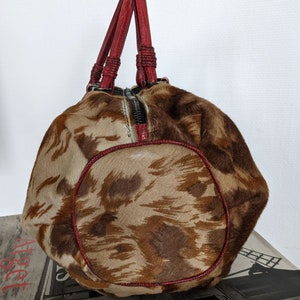 May include: A brown and tan patterned handbag with red leather handles. The bag has a zipper closure and a rounded shape.