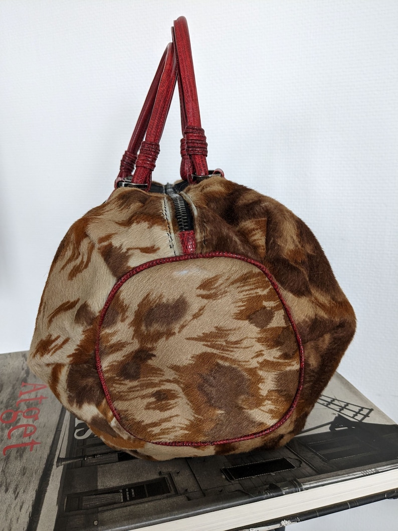 May include: A brown and tan patterned handbag with red leather handles. The bag has a zipper closure and a rounded shape.