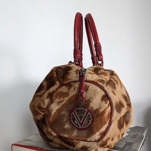 May include: A brown and tan handbag with a red leather trim and a large, round, silver-toned logo with rhinestones. The bag has two red leather handles.