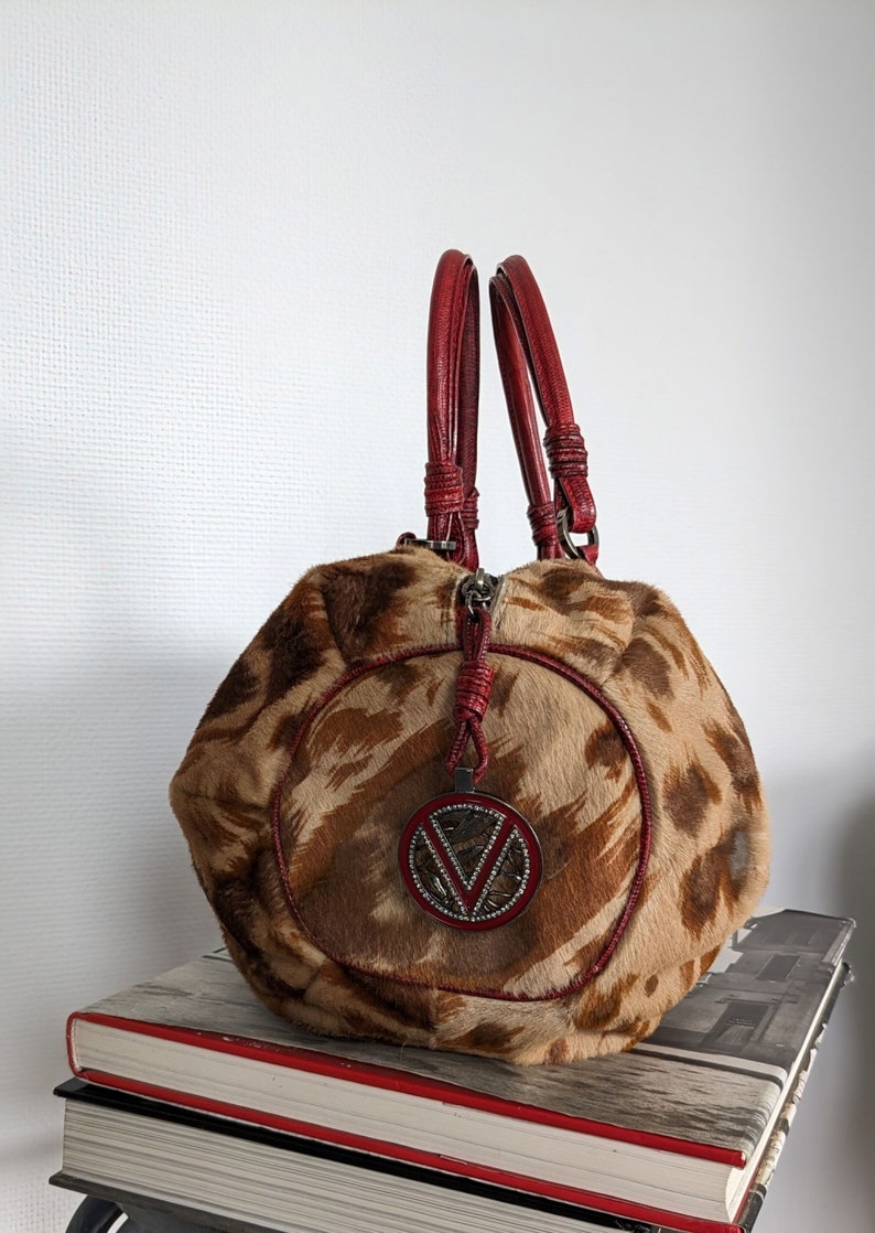 May include: A brown and tan handbag with a red leather trim and a large, round, silver-toned logo with rhinestones. The bag has two red leather handles.