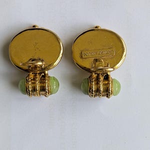 以下が含まれることがあります： ヴィンテージのジョルジオアルマーニのクリップオンイヤリングのペア。各イヤリングは、ブランド名と淡い緑色の楕円形の石が付いた、金色の円形のトップが特徴です。石は、金色の円筒形のデザインで上部に接続されています。