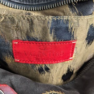 May include: Close-up of a red leather label with the text "VALENTINO GARAVANI" in white lettering. The label is attached to a brown fabric with a black and gold pattern.