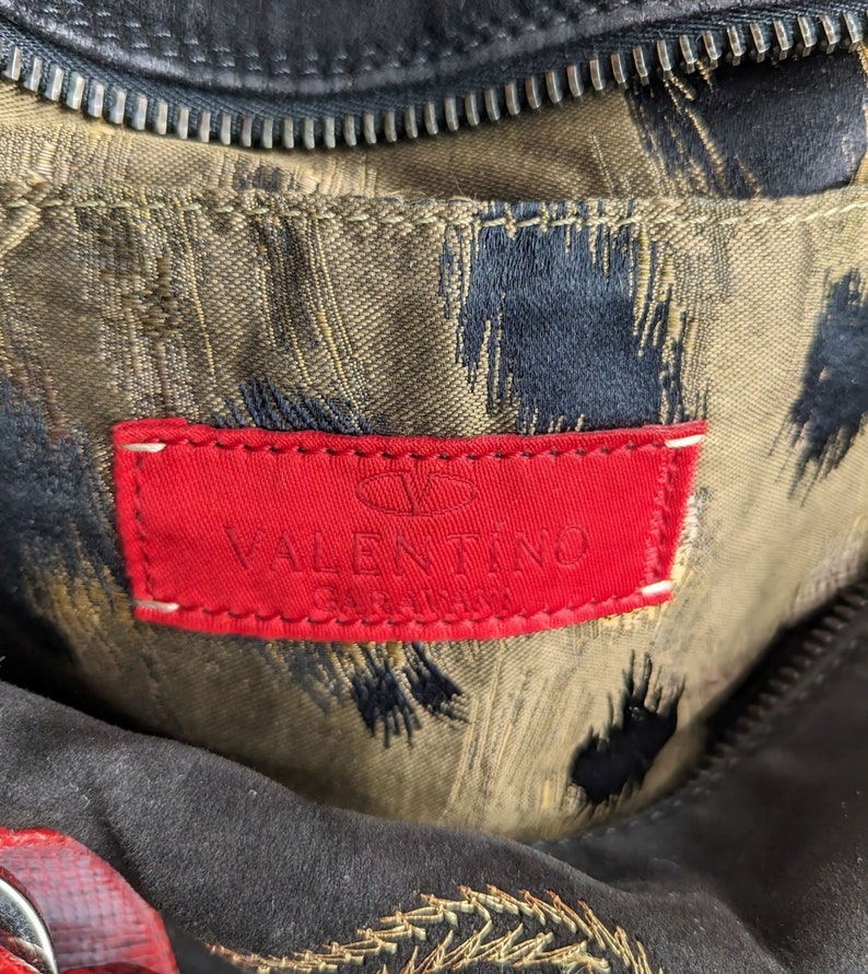 May include: Close-up of a red leather label with the text "VALENTINO GARAVANI" in white lettering. The label is attached to a brown fabric with a black and gold pattern.
