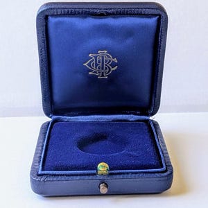 May include: Open navy blue jewelry box with a blue velvet interior. The box has a gold emblem on the inside lid and a gold clasp. The box is open, revealing a circular indentation in the base.