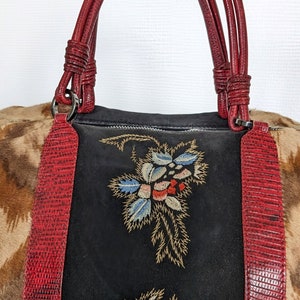 May include: A black suede handbag with red leather trim and embroidered floral designs. The bag has two top handles and a zipper closure.