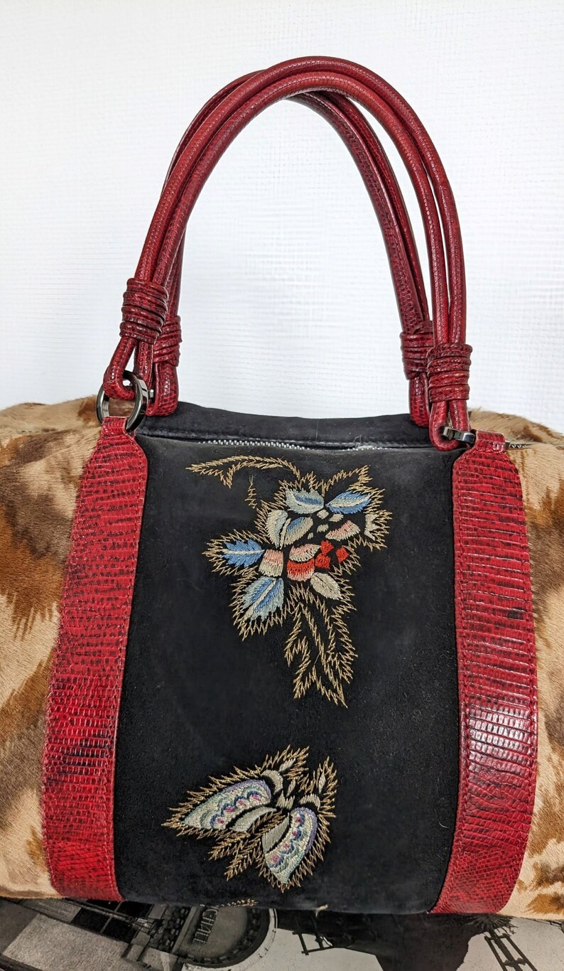 May include: A black suede handbag with red leather trim and embroidered floral designs. The bag has two top handles and a zipper closure.