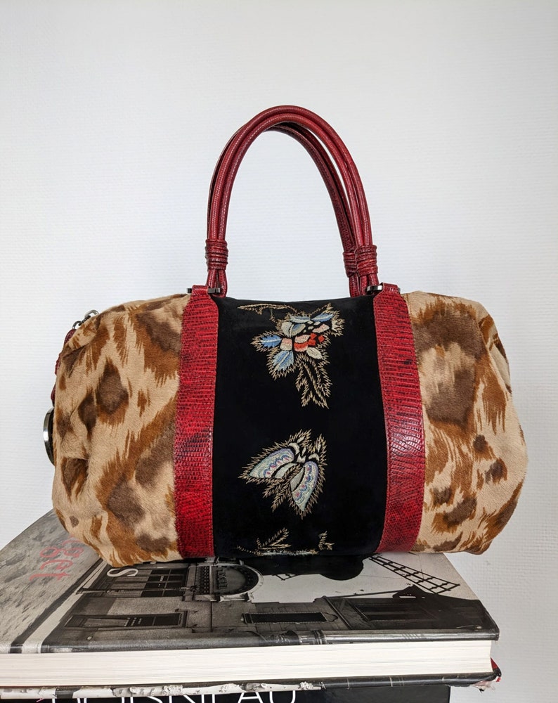 May include: A brown and tan leopard print handbag with red leather handles and trim. The bag has a black velvet panel with embroidered flowers in gold, blue, and red.