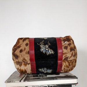 May include: A brown and tan leopard print handbag with red leather trim and embroidered floral details on the front. The bag is sitting on top of a stack of books.