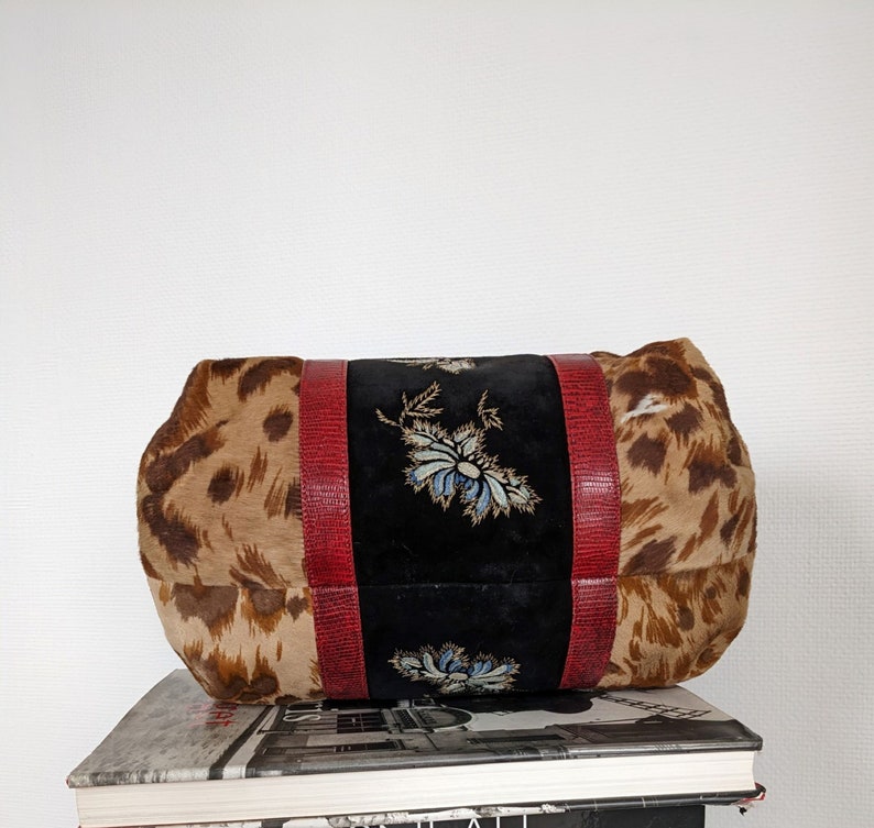 May include: A brown and tan leopard print handbag with red leather trim and embroidered floral details on the front. The bag is sitting on top of a stack of books.