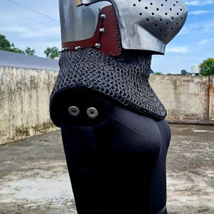 Op de afbeelding: Een middeleeuwse helm van gepolijst metaal, met een vizier met ventilatiegaten en een mali&euml;nkolder nekbescherming. De helm heeft een bruin lederen accent en wordt getoond op een zwarte mannequin.