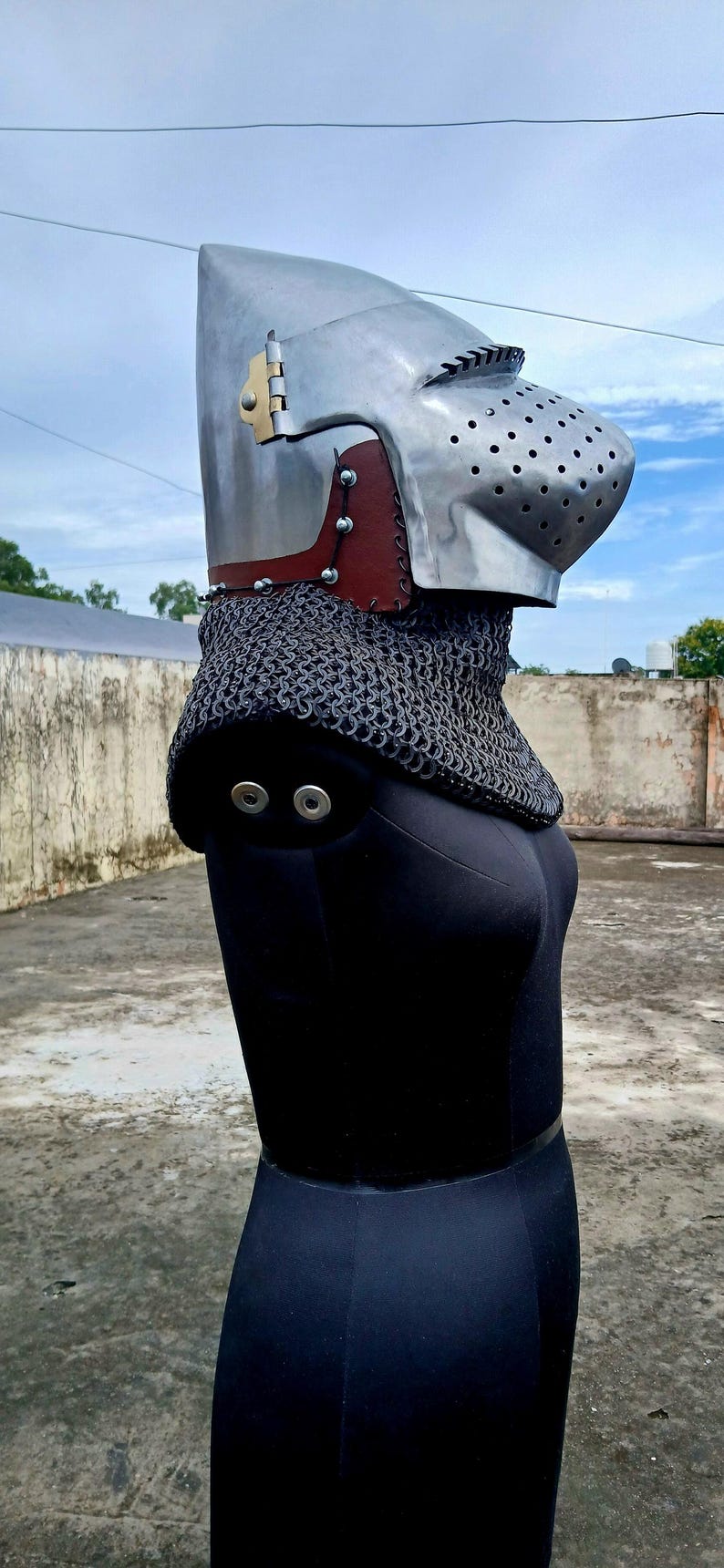 Op de afbeelding: Een middeleeuwse helm van gepolijst metaal, met een vizier met ventilatiegaten en een mali&euml;nkolder nekbescherming. De helm heeft een bruin lederen accent en wordt getoond op een zwarte mannequin.