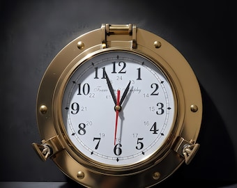 May include: A brass-colored, round clock with a white face and black numerals. The clock has a red second hand and is designed to resemble a ship's porthole. The clock face reads "Team & Supply".