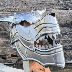 Puede incluir: Un casco de metal plateado con forma de cabeza de lobo, con dientes afilados y orejas puntiagudas. El casco tiene una superficie detallada y texturizada y está montado sobre un soporte de madera.
