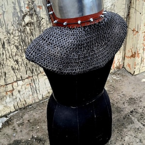 Op de afbeelding: Een middeleeuwse helm met een puntige bovenkant, gemaakt van glanzend metaal, staat op een zwarte mannequin. De helm heeft een bruine leren nekbeschermer en een mali&euml;nkolderkraag. Een goudkleurig metalen stuk is aan de voorkant bevestigd.