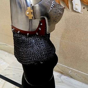 Op de afbeelding: Een middeleeuwse helm van gepolijst metaal, met een puntige bovenkant en een geperforeerde gezichtsbescherming. De helm is bevestigd aan een mali&euml;nkolder nekbescherming en een rode leren kraag. De helm wordt getoond op een zwarte vorm.