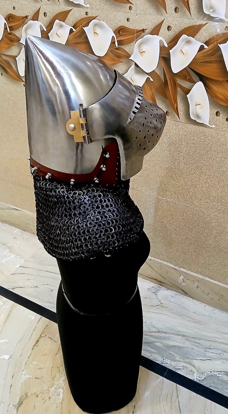Op de afbeelding: Een middeleeuwse helm van gepolijst metaal, met een puntige bovenkant en een geperforeerde gezichtsbescherming. De helm is bevestigd aan een mali&euml;nkolder nekbescherming en een rode leren kraag. De helm wordt getoond op een zwarte vorm.
