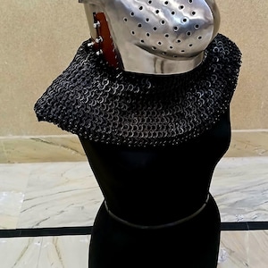 Op de afbeelding: Een gepolijste metalen helm met een vizier en een mali&euml;nkolder nekbescherming, weergegeven op een zwarte vorm. De helm heeft kleine gaatjes voor zicht en ventilatie. De mali&euml;nkolder is donker en gedetailleerd.