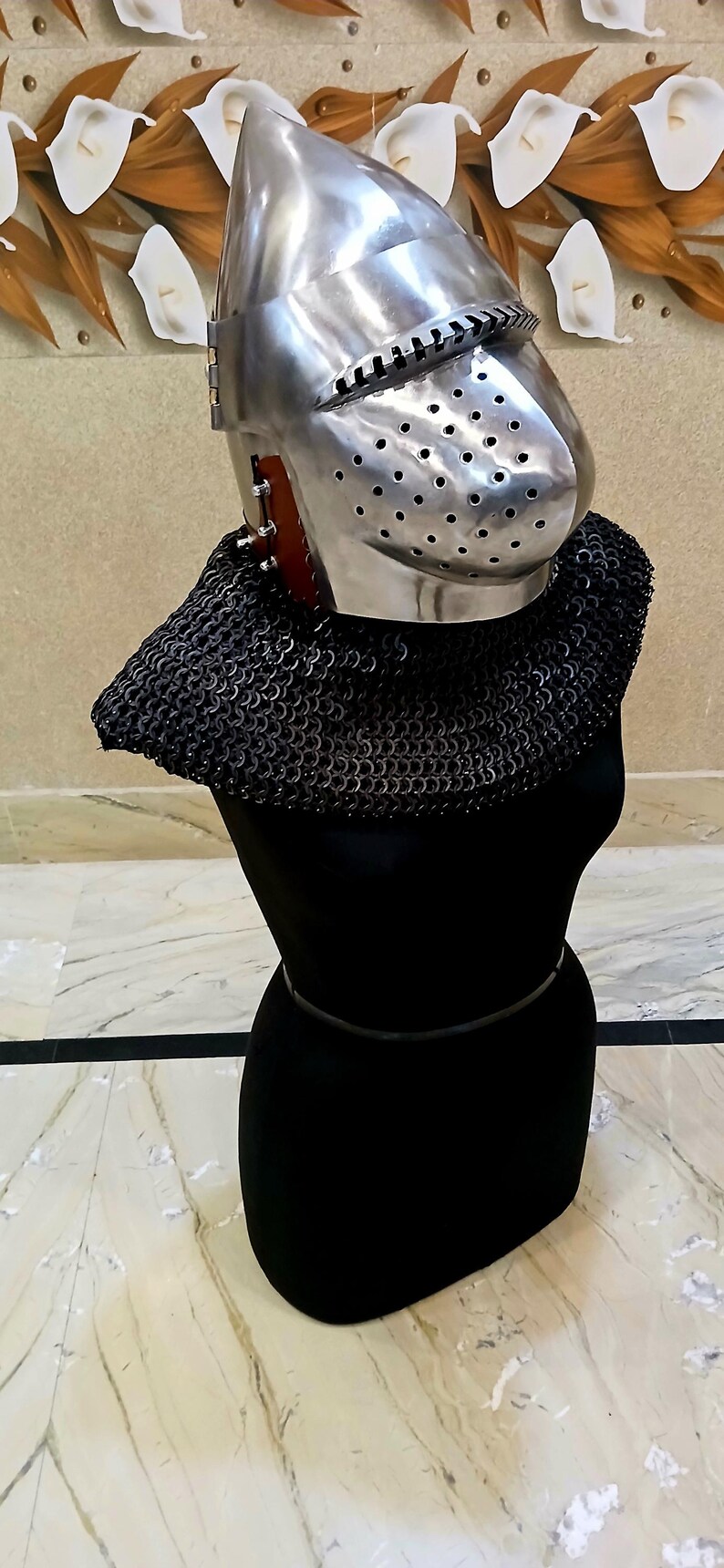 Op de afbeelding: Een gepolijste metalen helm met een vizier en een mali&euml;nkolder nekbescherming, weergegeven op een zwarte vorm. De helm heeft kleine gaatjes voor zicht en ventilatie. De mali&euml;nkolder is donker en gedetailleerd.