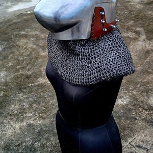 Op de afbeelding: Een middeleeuwse helm met vizier, gemaakt van gepolijst metaal. Het heeft een mali&euml;nkolder nekbescherming en is gemonteerd op een zwarte mannequin. De helm heeft leren en metalen details rond de nek.
