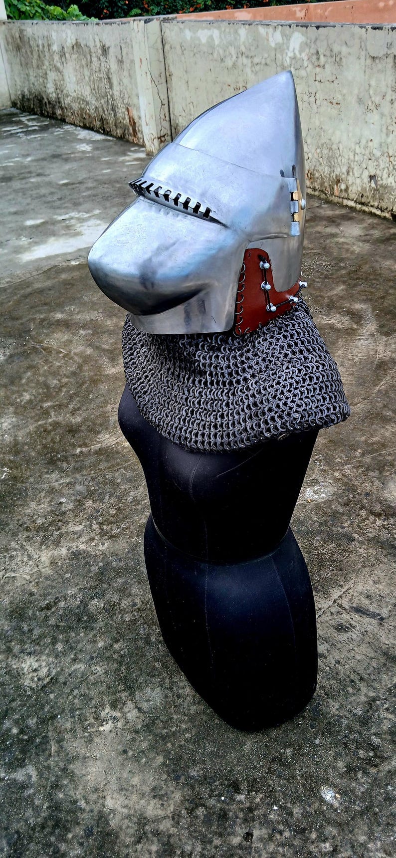 Op de afbeelding: Een middeleeuwse helm met vizier, gemaakt van gepolijst metaal. Het heeft een mali&euml;nkolder nekbescherming en is gemonteerd op een zwarte mannequin. De helm heeft leren en metalen details rond de nek.