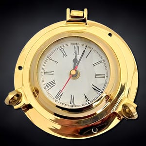 May include: A round, gold-colored clock with a white face and black Roman numerals. The clock has a red second hand and is encased in a brass-like frame with a porthole design. The clock's hands point to approximately 12:00. The clock is a nautical-themed decorative item.