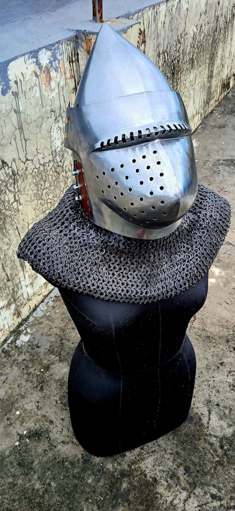 Op de afbeelding: Een middeleeuwse helm van glanzend metaal met een puntige bovenkant en een vizier met kleine gaatjes. Wordt geleverd met een mali&euml;nkolder kraag, gepresenteerd op een zwarte mannequin. Ideaal voor re-enactment.