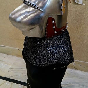 Op de afbeelding: Een gepolijste metalen helm met een puntig vizier en een mali&euml;nkolder nekbescherming. De helm heeft een rode leren afwerking en wordt getoond op een zwarte mannequin. De achtergrond heeft een bloemen wandontwerp.