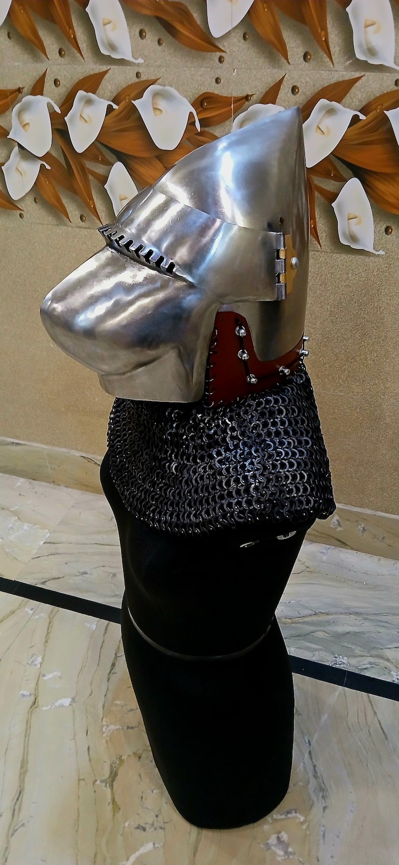 Op de afbeelding: Een gepolijste metalen helm met een puntig vizier en een mali&euml;nkolder nekbescherming. De helm heeft een rode leren afwerking en wordt getoond op een zwarte mannequin. De achtergrond heeft een bloemen wandontwerp.