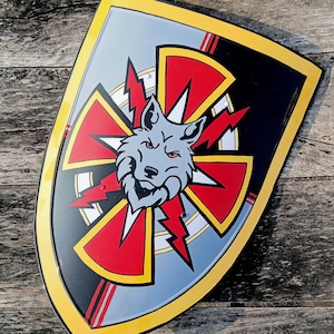 Puede incluir: Un emblema en forma de escudo con la cabeza de un lobo en el centro, rodeada por un diseño rojo y blanco con rayos. El escudo tiene un fondo negro con un borde amarillo y está sobre una superficie de madera.