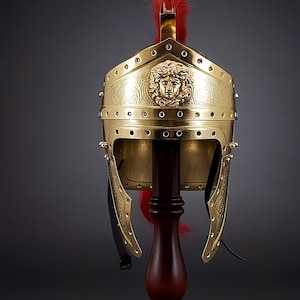 May include: A gold-colored Roman helmet with a red plume, displayed on a wooden stand. The helmet features a detailed face design and decorative rivets. The helmet is a historical replica.