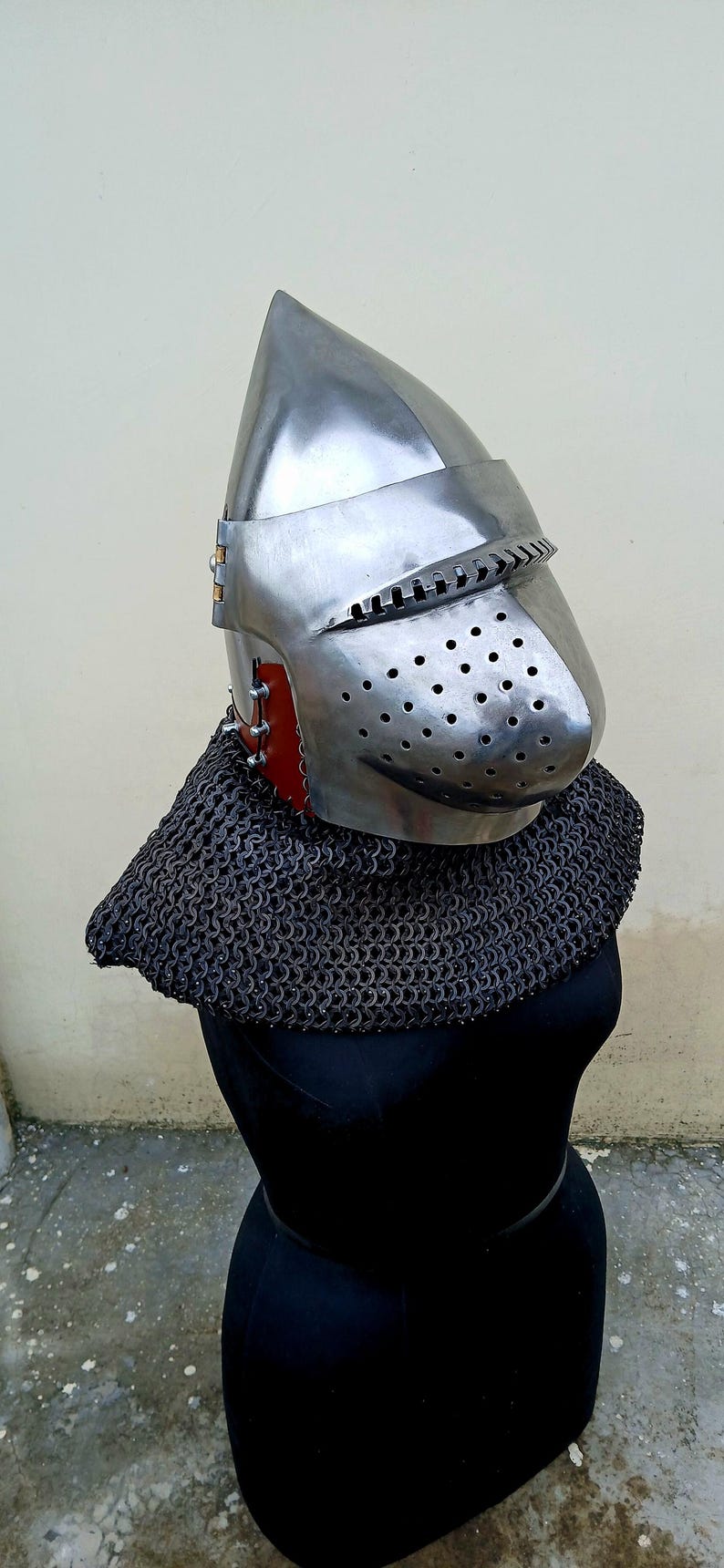Op de afbeelding: Een gepolijste middeleeuwse stalen helm met een vizier en ventilatiegaten. De helm wordt gecombineerd met een mali&euml;nkolder, gepresenteerd op een zwarte mannequin. Het ontwerp doet denken aan historische harnassen.