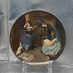 Puede incluir: Plato decorativo con una ilustración detallada de un hombre leyendo a una niña, con un perro y libros. La obra de arte está en tonos cálidos, con una estética vintage. El plato se exhibe en un soporte transparente.
