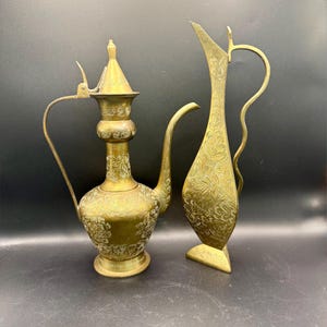 May include: Two antique brass pitchers with intricate floral patterns. One pitcher has a long, curved spout and a hinged lid. The other is tall and slender with a curved handle. Both are displayed against a dark background.