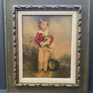 Puede incluir: Retrato enmarcado antiguo de un niño pequeño sosteniendo un perro pequeño y esponjoso. El niño lleva una chaqueta roja, pantalones color canela y una camisa blanca con cuello. Un sombrero de copa negro descansa en el suelo cerca del niño. El marco es de color verde desgastado.