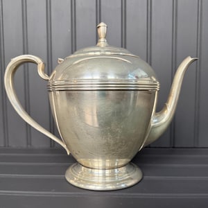 Puede incluir: Tetera plateada con asa y boquilla curvadas. La tetera tiene una tapa con un pequeño adorno y se asienta sobre una base redonda. El diseño es clásico y elegante.