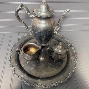 May include: A vintage silver tea set, including a teapot, creamer, sugar bowl, and a round serving tray. The set has ornate detailing and a tarnished finish, suggesting age and use. The tray has a decorative floral border.