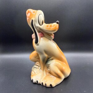 May include: A ceramic figurine of Pluto, the Disney cartoon dog, in a seated position. The figurine features a tan and orange color scheme with black accents on the nose, ears, and paws. A pink bow tie is around the neck.
