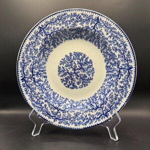 May include: A white and blue ceramic bowl with a detailed tree branch pattern. The bowl has a central design and a repeating pattern around the rim. The bowl is displayed on a clear stand.