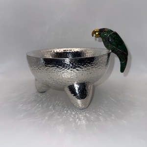 Könnte beinhalten: Eine silberfarbene, gehämmerte Metallschale mit drei abgerundeten Füßen und einer dekorativen Papageienfigur am Rand. Der Papagei ist aus grünem, steinartigem Material gefertigt und besitzt einen goldfarbenen Schnabel sowie ein kleines rotes Auge.