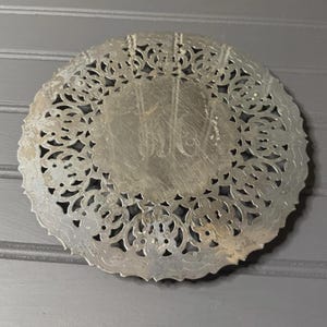 以下が含まれることがあります： 銀色の丸い装飾プレート。縁には複雑な透かし彫りのデザインが施されています。中央にはエッチングされたモノグラムがあります。このプレートはおそらく金属製で、ヴィンテージまたはアンティークの美しさを持っています。