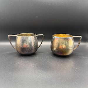 May include: Two silver-toned, small, handled serving pieces. One is a creamer with a spout, and the other is a sugar bowl with two handles. Both have a rounded shape and a tarnished appearance, suggesting age.