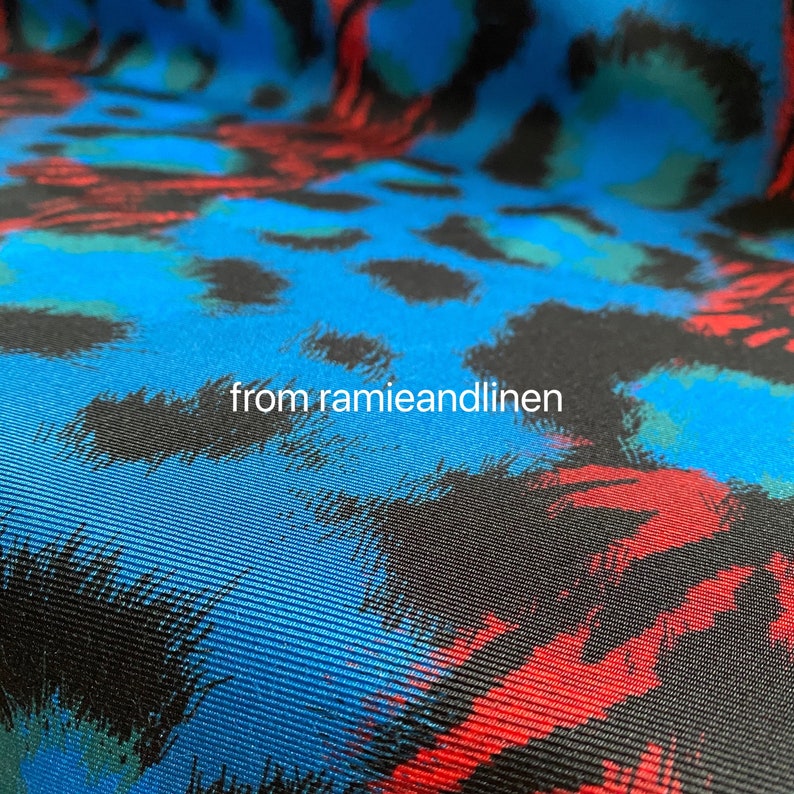 May include: Close-up of a blue fabric with a black, red, and green animal print. The fabric has a textured, woven appearance.