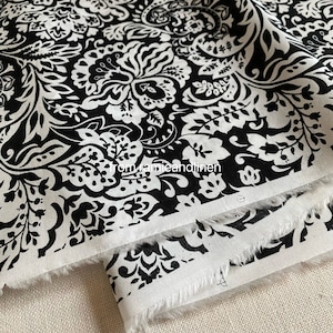 May include: Black and white floral fabric with a damask pattern. The fabric features a repeating design of stylized flowers and leaves. The edges of the fabric are frayed. The text "from jamieandlinen" is visible.