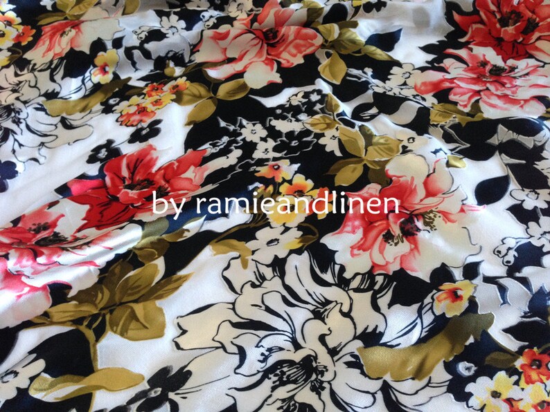 May include: White fabric with a black background and a large floral print in shades of red, pink, white, and yellow. The fabric has a satin-like sheen. The text "by ramieandlinen" is printed on the fabric.