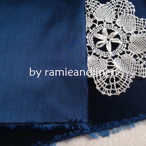 May include: A close-up of a dark blue fabric with a white crocheted lace doily on top. The fabric has a fringed edge.